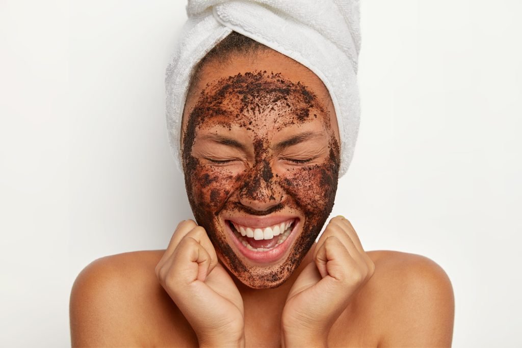 donna con turbante di spugna bianco che ha sulla faccia uno scrub per l'esfoliazione viso e sorride portandoi i pugni verso il volto