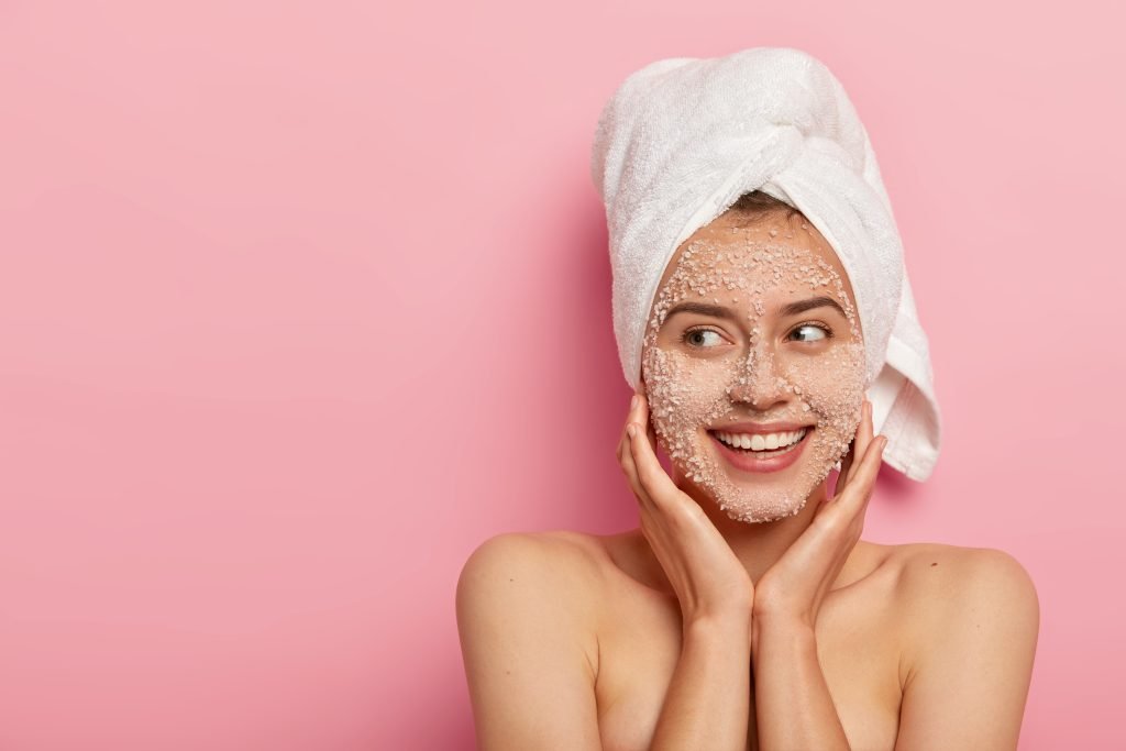ESFOLIAZIONE VISO RAGAZZA CON ASCIUGAMANO bianco IN TESTA E SCRUB per esfoliazione VISO su parete con SFONDO ROSA