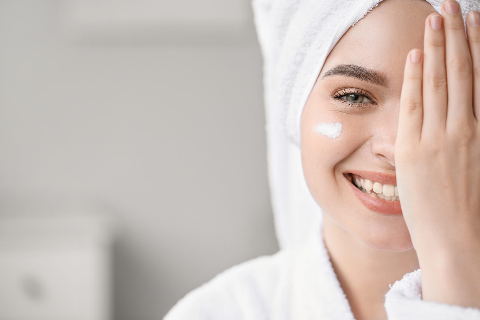 concetto di routine skincare mare - ragazza ritratta con accappatoio e asciugamano a turbante sul capo, sorride e si copre meta viso con la mano