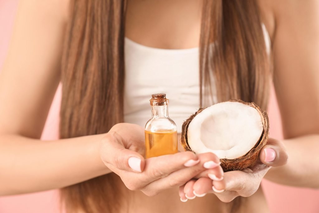 primo piano di mezzo busto di donna con capelli lunghi che tiene posati sulle mani una boccetta d'olio e un cocco a metà. Concetto cura dei capelli e hair oiling 
