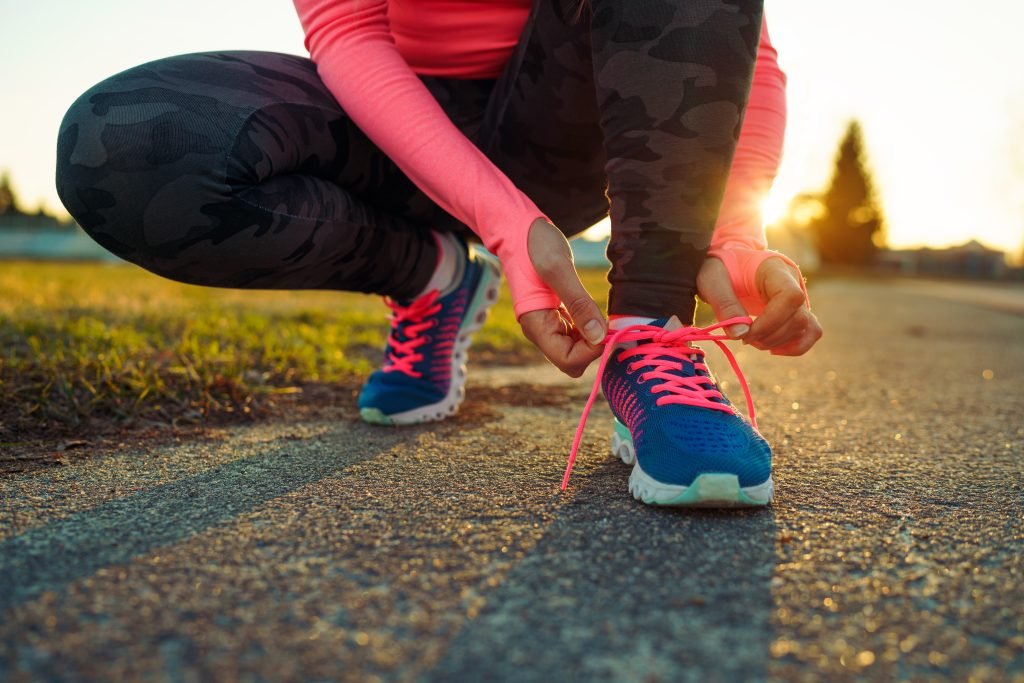 primo piano su mezzo busto inferiore di ragazza accosciata che si allaccia con il sole che tramonta alle spalle la scarpa da camminata per fare i 10000 passi al giorno
