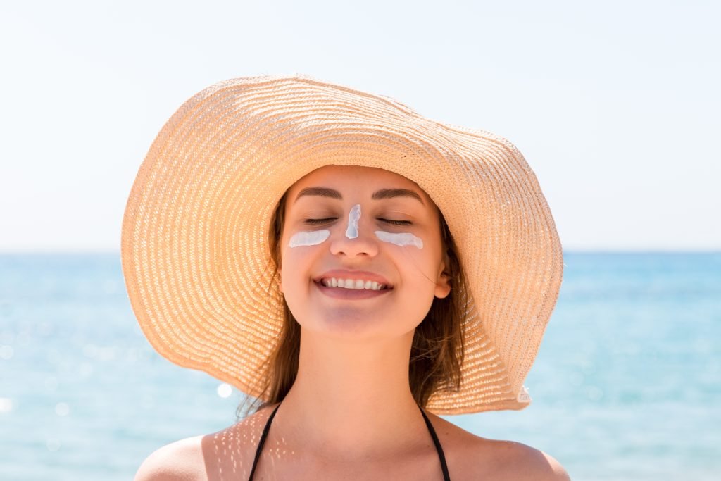 primo piano di giovane donna al mare con cappello di paglia, occhi chiusi mentre sorride . Crema solare spalmata su zigomi e naso .