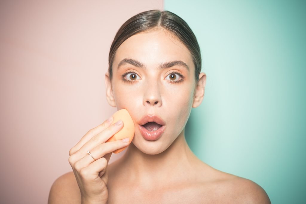 ragazza stupita si trucca con una spugnetta a forma di uovo per ottenere un look naturale e luminoso con primer illuminanti e correttore 