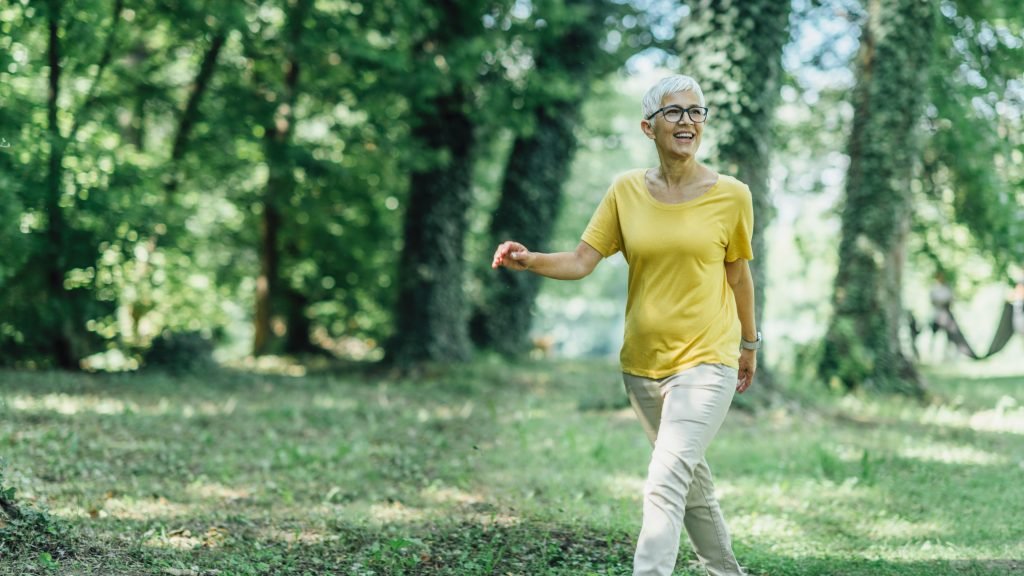 donna di mezza eta che cammina in un bosco con maglia gialla e pantalone chiaro capelli corti bianchi e occhiali per esprimere il concetti dei benefici dei 10000 passi al giorno in menopausa