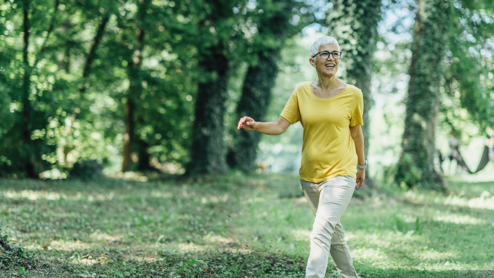 donna di mezza eta che cammina in un bosco con maglia gialla e pantalone chiaro capelli corti bianchi e occhiali benefici dei 10000 passi al giorno in menopausa PER CONTRASTARE sarcopenia e osteoporosi 