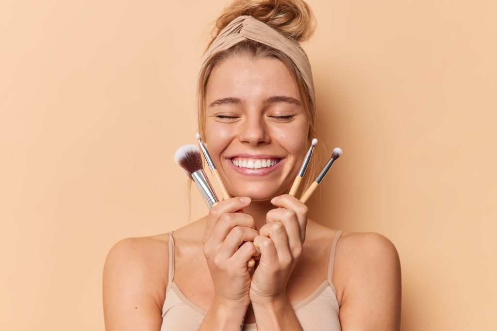 ragazza su sfondo arancione pallido ritratta a mezzo busto con capelli raccolti con una fascia beige che sorride e tiene gli occhi chiusi mentre porta al viso dei pennelli da makeup per i primer illuminanti