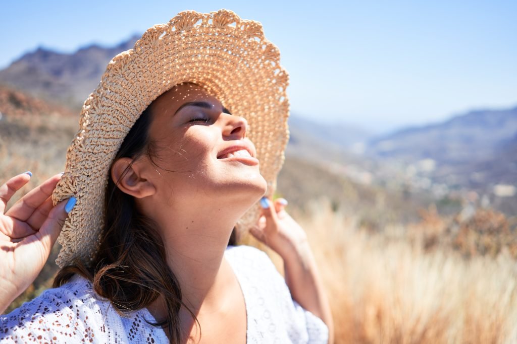 ragazza in primo piano con cappello di paglia tenito con le mani con smalto azzurro e vestito in pizzo bianco che espone il viso al sole sorridendo e con occhi chiusi con makeup leggero e naturale e protezione solare da usare sopra il trucco