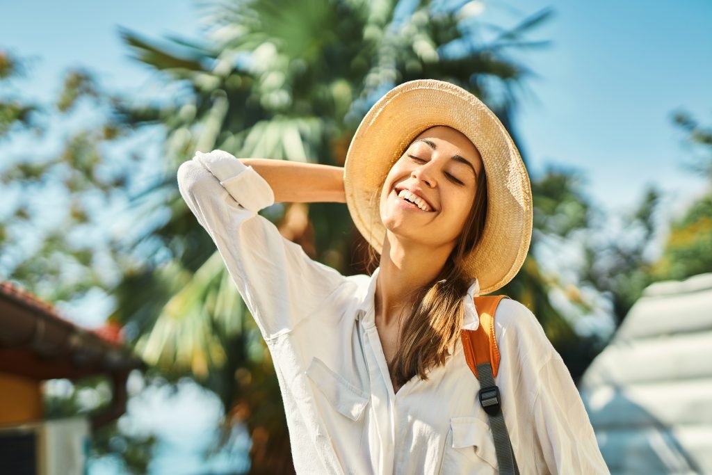 ragazza che sorride e volge il viso al sole tnendo occhi chiusi vestita con cappello di paglia e camicia bianca con makeup leggero e protezione solare da usare sopra il trucco