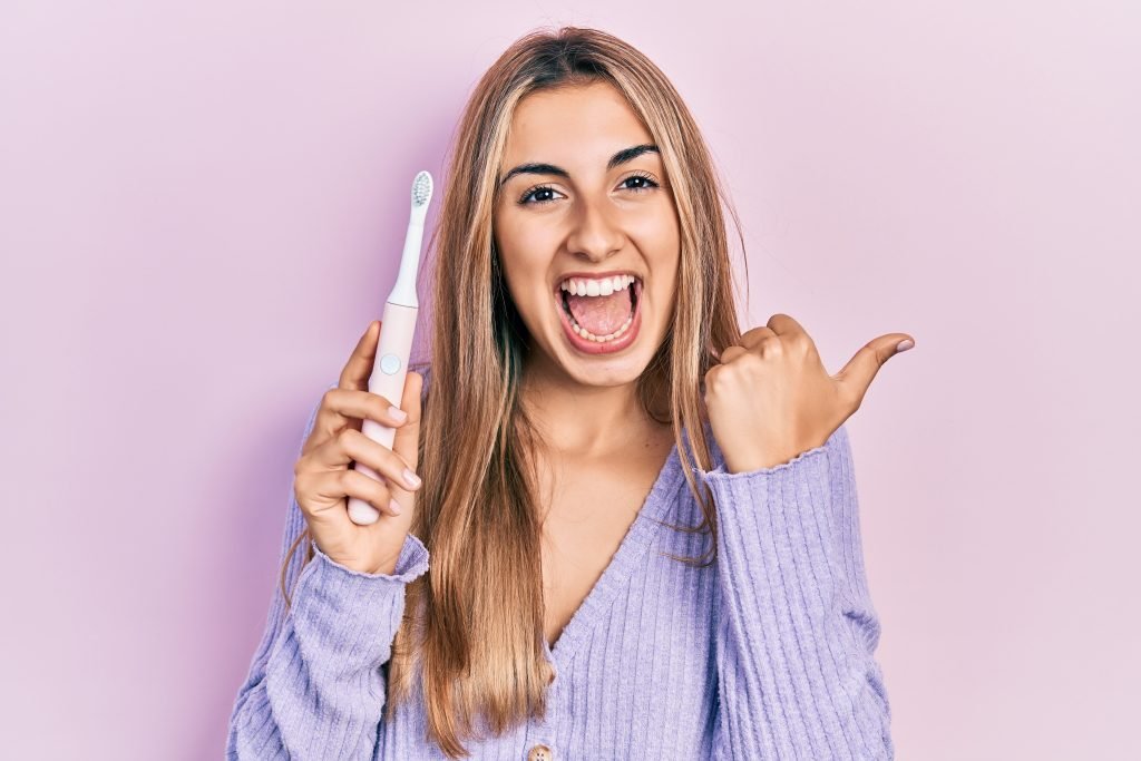 ragazza bionda vestita di viola che sorride entusiasta e fa ok con una mano mentre con l'altra tiene in mano lo spazzolino elettrico su sfondo rosa 