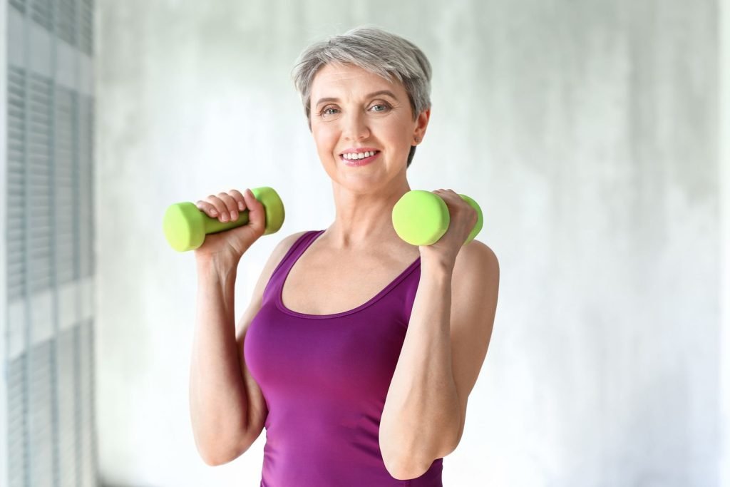 concetto di attività fisica per prevenire l'osteoporosi donna matura che si allena con i pesi con canotta viola e pesi gialli guarda in camera e sorride sfondo sfumato neutro