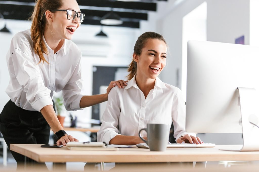 concetto del come essere più produttivi : colleghe in ufficio che sorridono mentre entrambe guardano lo schermo di un pc alla scrivania con camicia bianca 