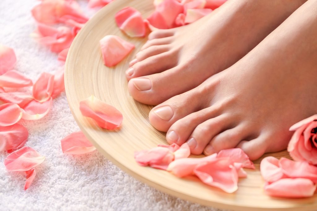 primo piano di piedi di donn posati su un vassoio in legno su cui ci sono petali di rosa rosa. Concetto di cura di sè e di pedicure fai da te