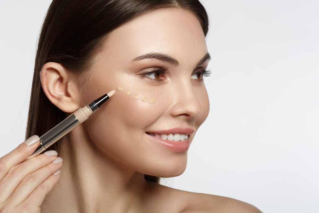 Joyful young woman using concealer and smiling. She is holding special brush near her face. Drops of foundation are on cheek. Isolated and copy space