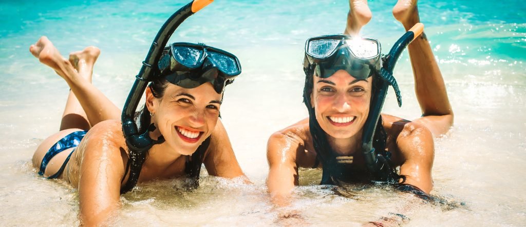 primo piano di ragazze sorridenti sdraiate sul bagnasciuga al mare con indosso maschera (sulla fronte ) e boccaglio per fare snorkeling - concetto di idee su cosa mettere nella borsa mare