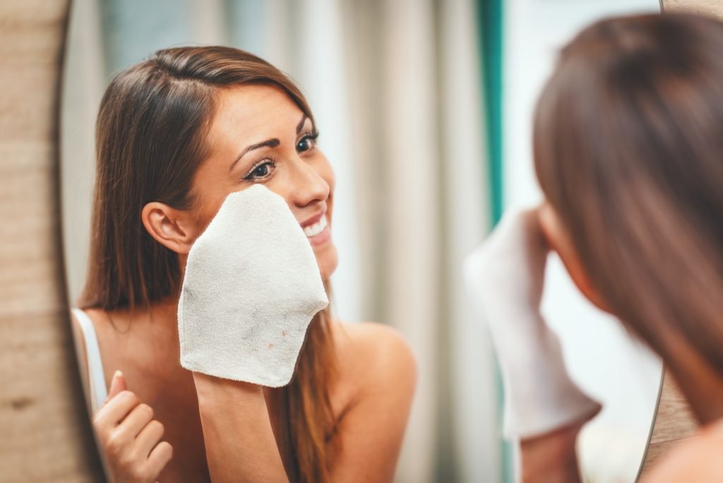 Giovane donna sorridente rimuover il makeup nella prima fase della doppia detersione con il guanto in microfibra davanti allo specchio 