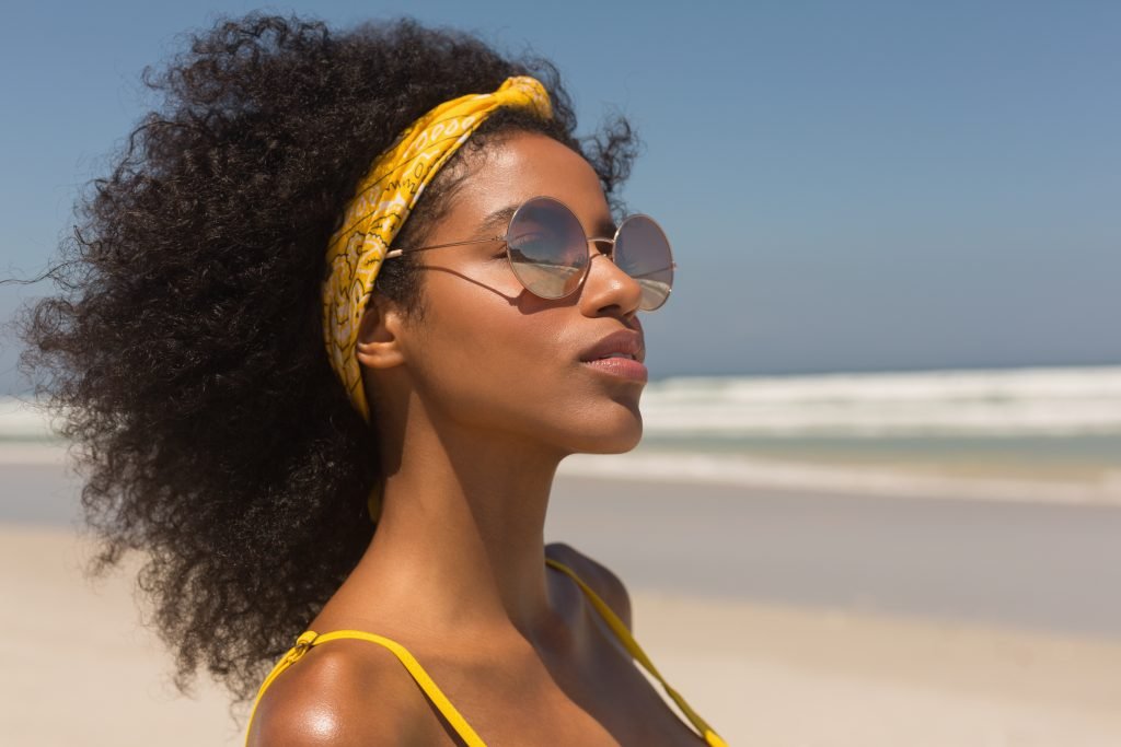 primo piano di donna afro con occhiali da sole fascia capelli e costume giallo. Concetto di cura capelli al mare ricci