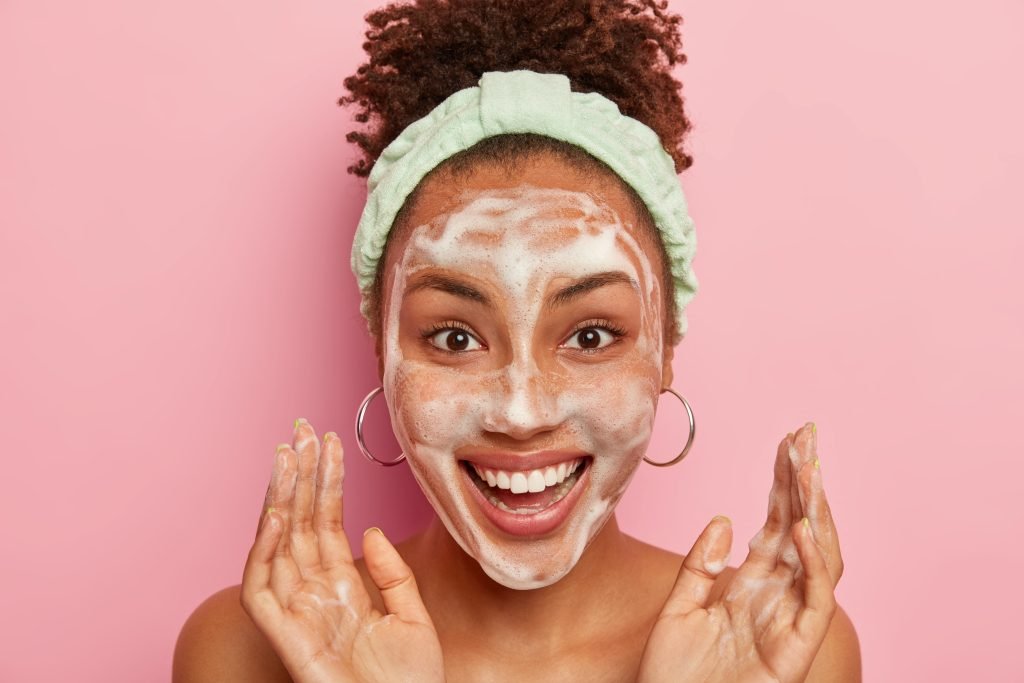 PRIMO PIANO DI RAGAZZA AFRO AMERICANA CON CAPELLI RACCOLTI DA FASCIA PER CAPELLI VERDE CHIARO E ORECCHINI A CERCHIO. HA LA FACCIA SORRIDENTE E SORPRESA E SORRIDE MENTRE FA DETERSIONE DEL VISO . HA SCIUMA DEL SERTEGENTE SUL VISO SFONDO ROSA 