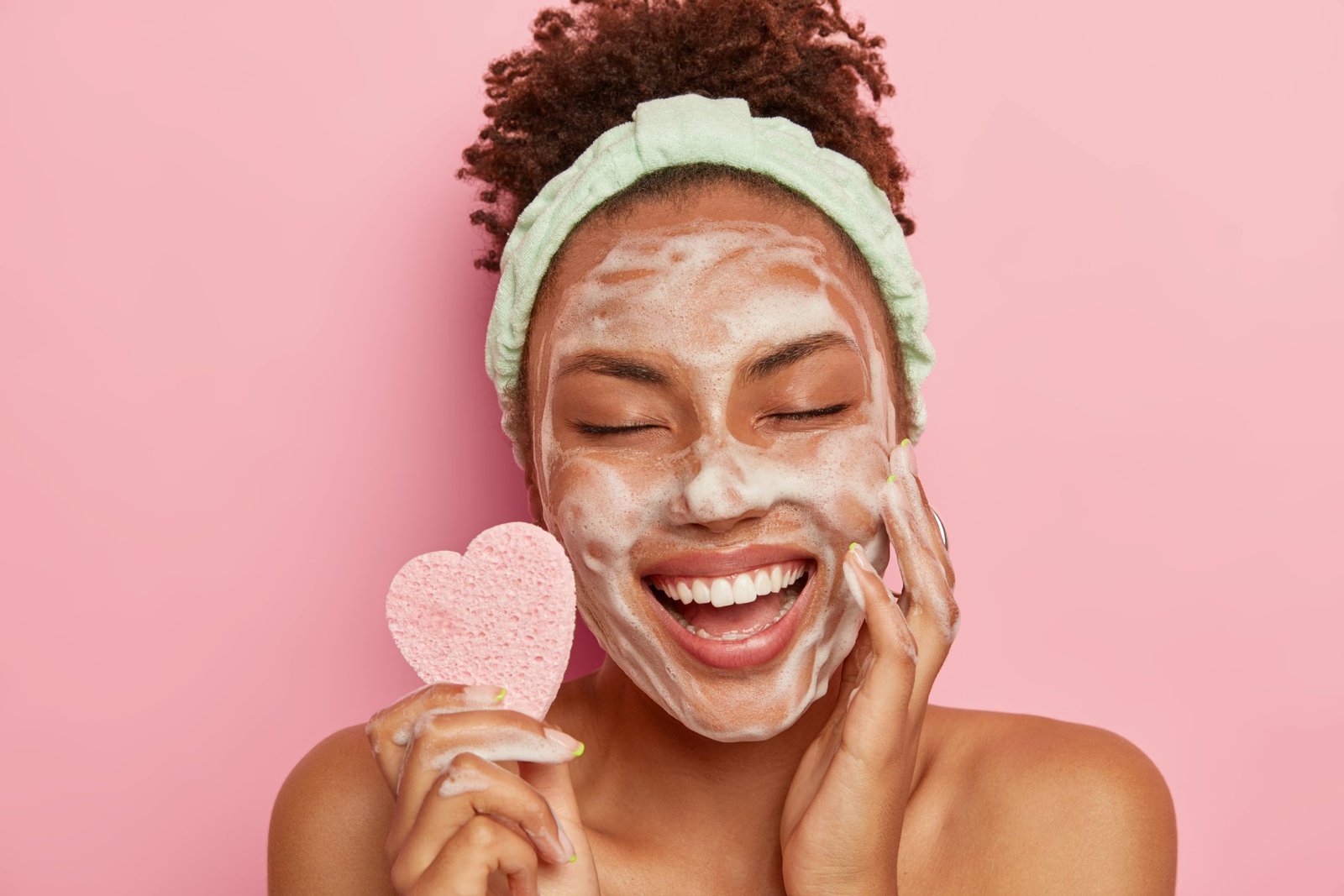 Modella rilassata e gioiosa dalla pelle scura si lava il viso con bolle di sapone, si gode una sessione di coccole, tiene gli occhi chiusi per non provare piacere, tiene in mano una spugna cosmetica, si prende cura del suo corpo, sta nuda in casa. concetto di routine skincare mare