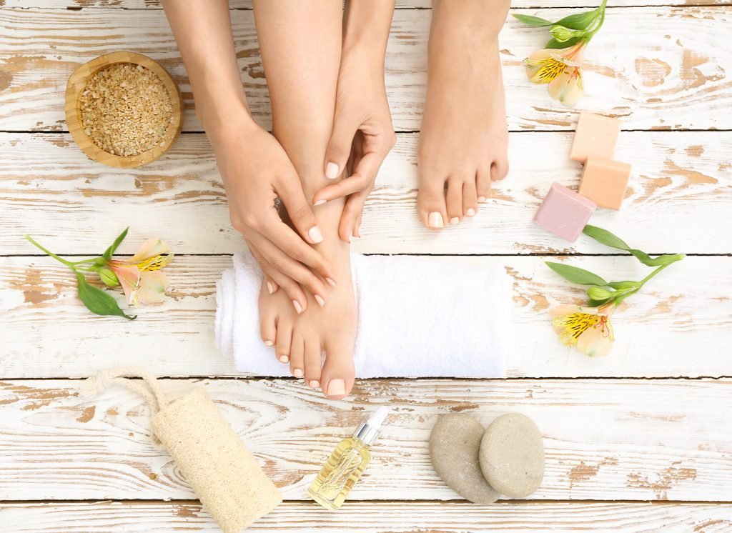 primo piano con immagine scattata dall'alto di peidi di donna su pavimento di legno decorato con alcuni fiori e con alcuni prodotti necessari per una pedicure fai da te casalinga