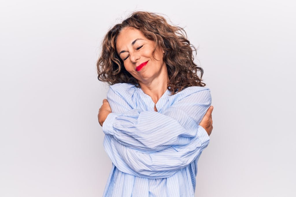 Bella donna bruna di mezza età che indossa una camicia a righe in piedi su uno sfondo bianco, che si abbraccia felice e positiva, sorridendo e sicura di sé. Amore per se stessi e cura di sé.