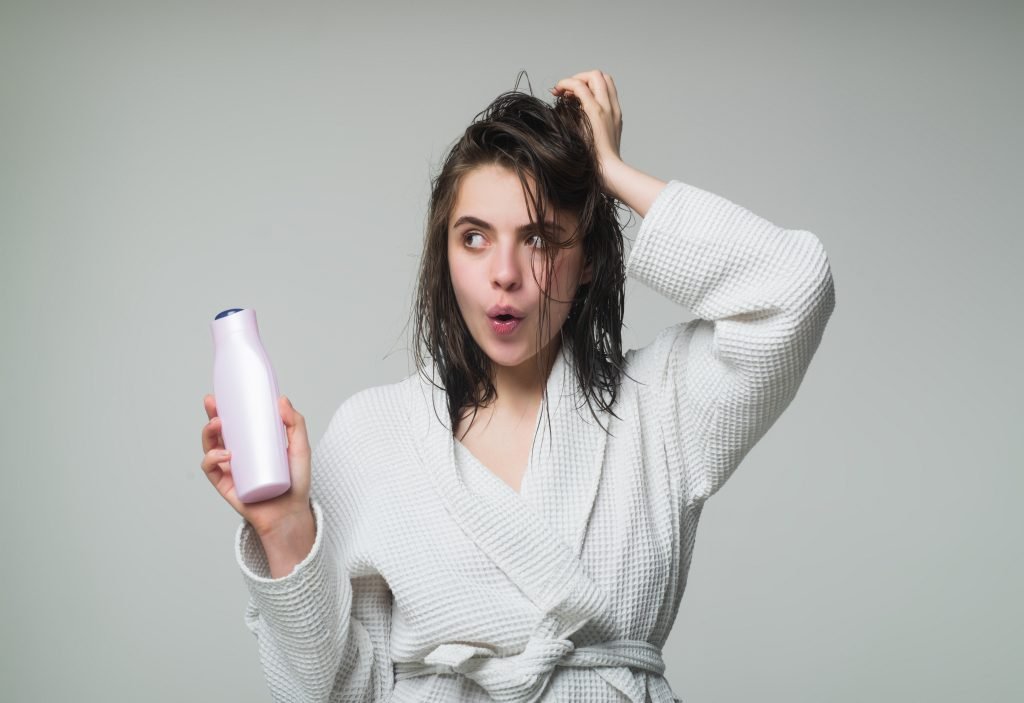 Donna che si tocca i capelli in accapatoio bianco dopo la doccia e tiene in mano una bottiglia di shampoo e balsamo. Sfondo neutro grigio . espressione stupita o perplessa in merito al risultato sui suoi capelli