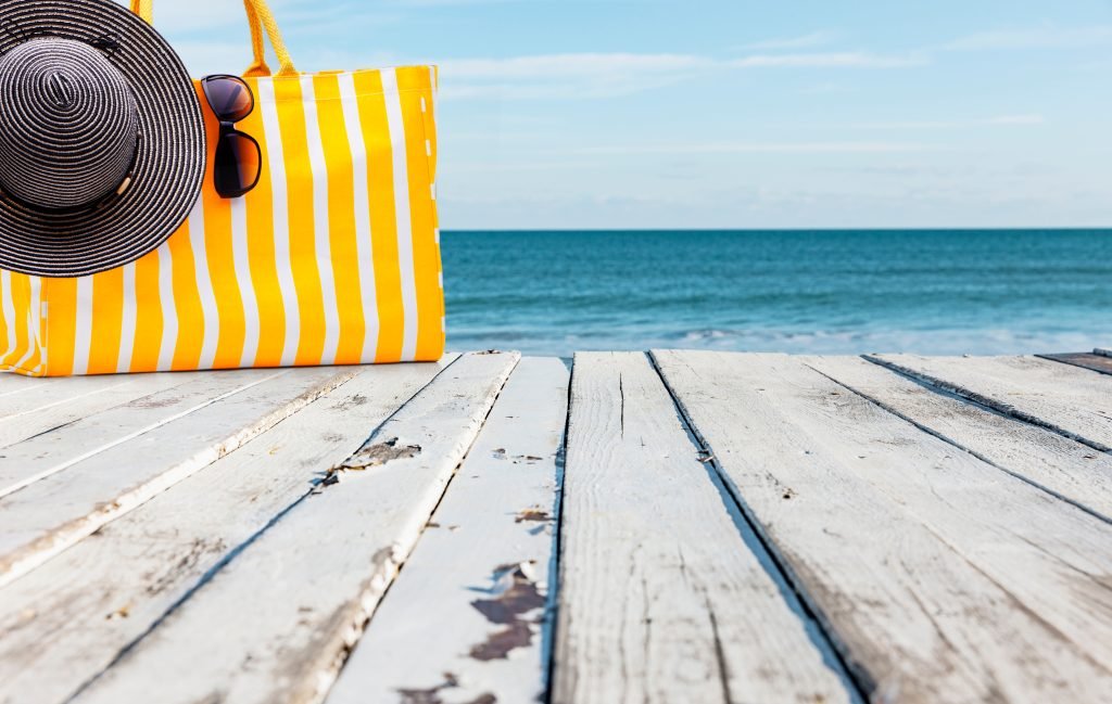 panorama del mare fotografato da una piattaforma in legno su cui èp posata una borsa mare gialla a righe bianche con un cappello di paglia nero e degli occhiali da sole. Concetto di vacanze