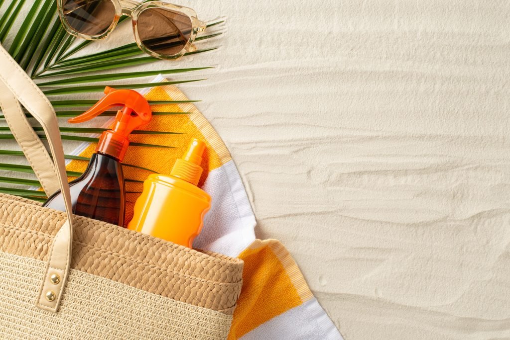 Concetto di cura capelli al mare. Foto dall'alto di una borsa di vimini con spray solari capelli , occhiali da sole e foglie di palma su un telo da spiaggia su uno sfondo di sabbia isolata con spazio per il testo.