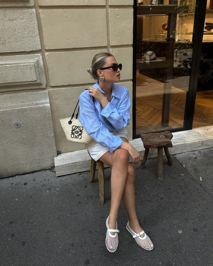 ragazza con capelli raccolti e occhiali da sole seduta fuori da un bar con camicia azzurra e pantaloncino chiaro e le ballerine bianche in rete di Alaia 