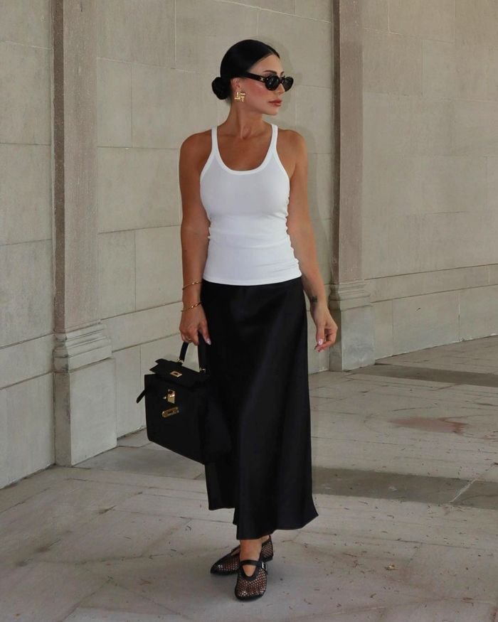 giovane donna con capelli raccolti canottiera bianca e gonna lunga nera che passeggia per strada con borsa nera e ballerine in rete economiche 