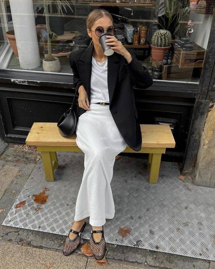 donna seduta fuori da un bar che bece un te vestita con outfit bianco blazer nero e ballerine in rete di Alaia 