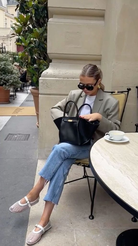 ragazza seduta al tavolo di un bar vestita con blazer beige , jeans e ballerine in rete economiche bianche che cerca qualcosa nella borsa