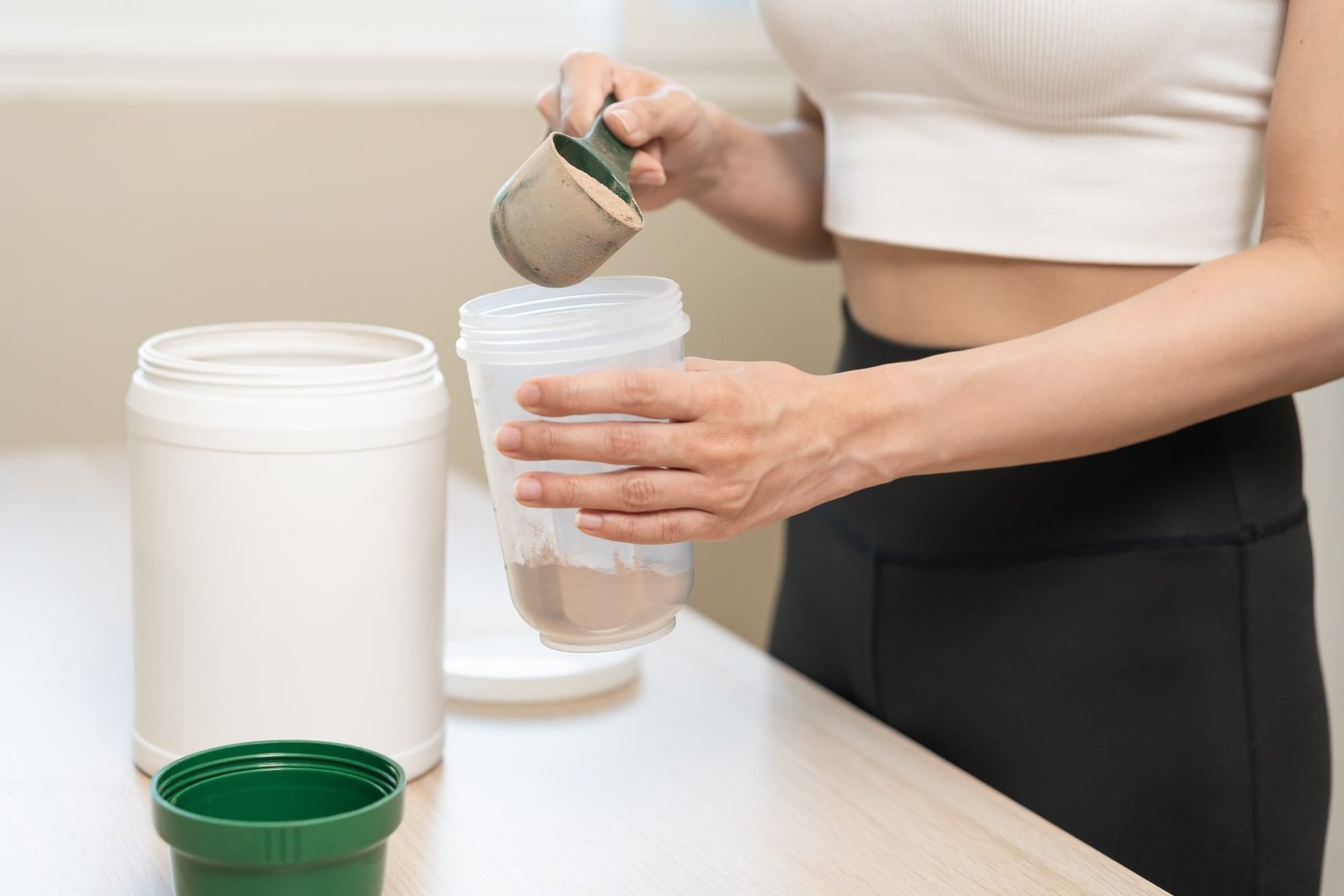 mezzo busto senza viso di giovane donna in abbigliamento sportivo, mano che tiene un misurino per preparare le proteine nella bottiglia da shakerare, integratore per i muscoli dopo l'allenamento a casa. Cura del corpo sana.