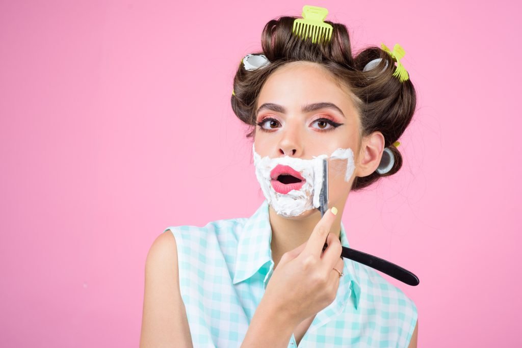 Ragazza pin-up con BIGODINI IN TESTa e trucco. stile retrò mentre si rade con schiuma e lametta. Concetto di dermaplaning pro e contro