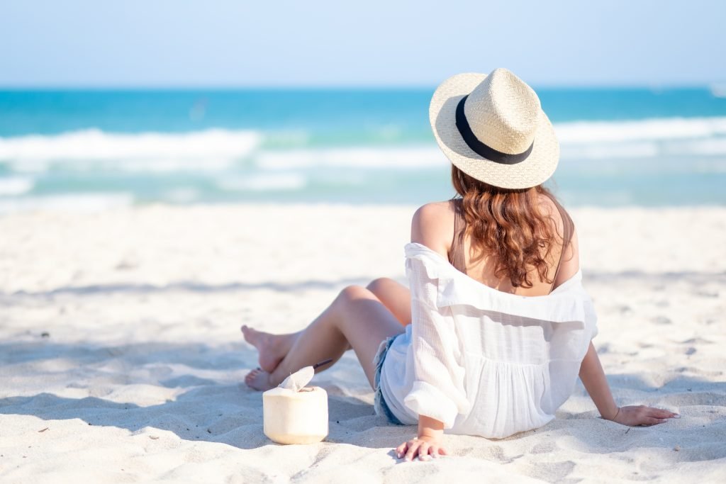 Ritratto di una bella donna di spalle in abito e cappello di paglia bianchi che si siede e beve succo di cocco sulla spiaggia metre osserva il mare