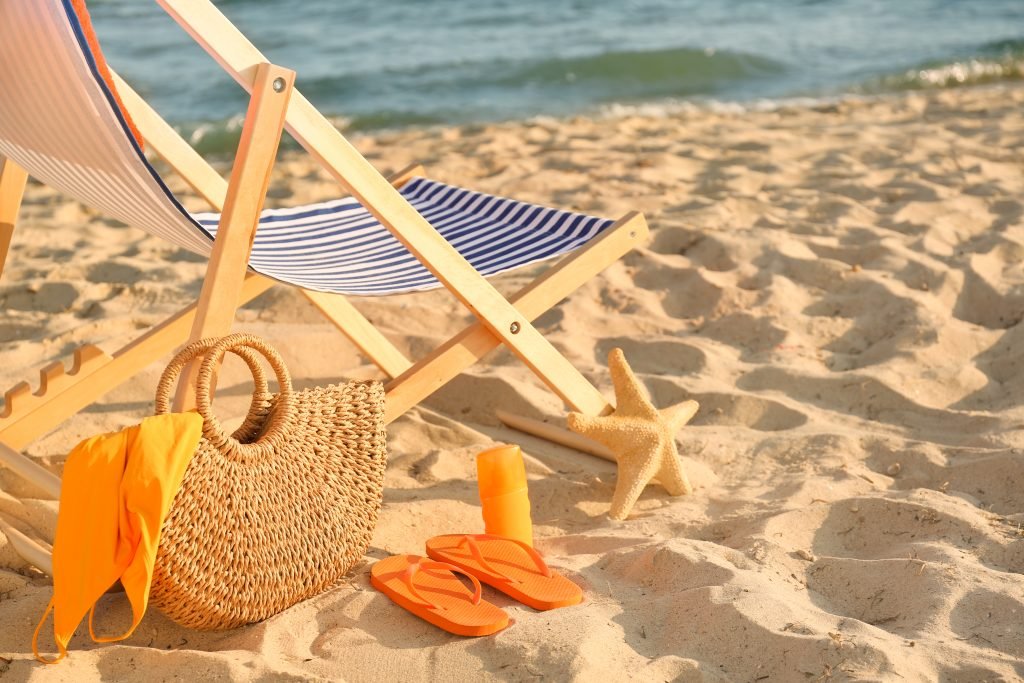 sdraio a righe sulla sabbia. sfondo del mare . accanto alla sdraio borsa in paglia costume e ciabatte - concetto di idee su cosa mettere nella borsa mare