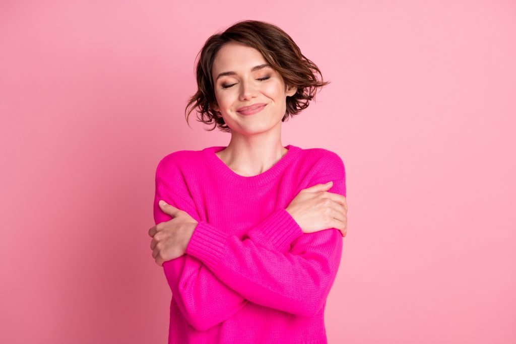 Foto di una donna attraente e gioiosa, di buon umore, con le mani che si abbracciano e le spalle che si godono la gioia, il calore del panno morbido e gli occhi chiusi, indossando un maglione casual fuxia luminoso isolato su sfondo rosa pastello.
