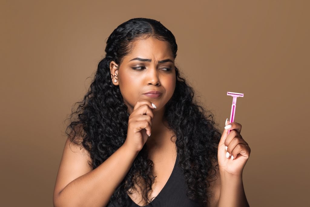 Donna latina curvy pensierosa che guarda il rasoio di sicurezza e si tocca il mento, in piedi e in posa isolata su sfondo marrone da studio. Concetto di dermaplaning pro e contro 