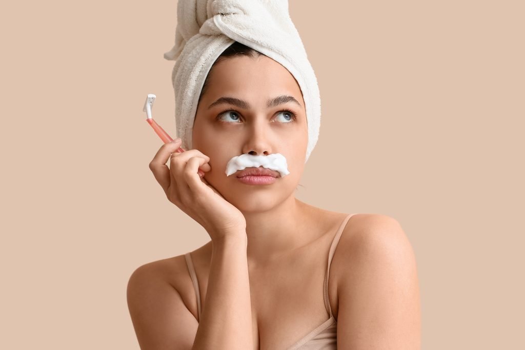 Bella giovane donna perplessa e pensierosa con asciugamano in testa e con rasoio e schiuma da barba su labbra superiore su sfondo beige. Concetto di dermaplaning pro e contro 