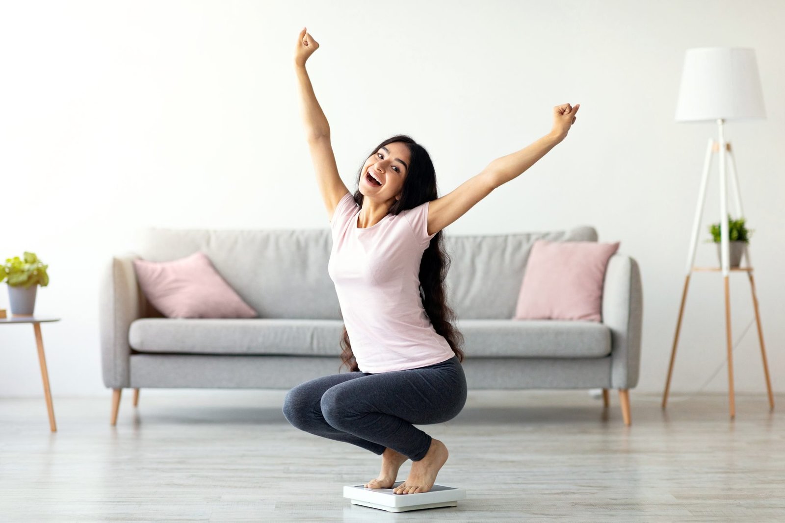 Donna entusiasta seduta sulla bilancia, alza le mani, felice dei risultati del suo programma di dimagrimento e allenamento a casa. Concetto di motivazione per sport e dieta 