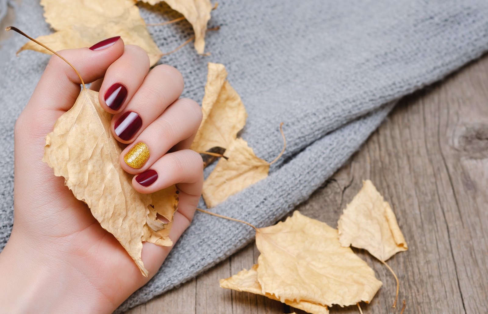 Mano femminile con unghie decorate di colore rosso scuro che regge foglie gialle.