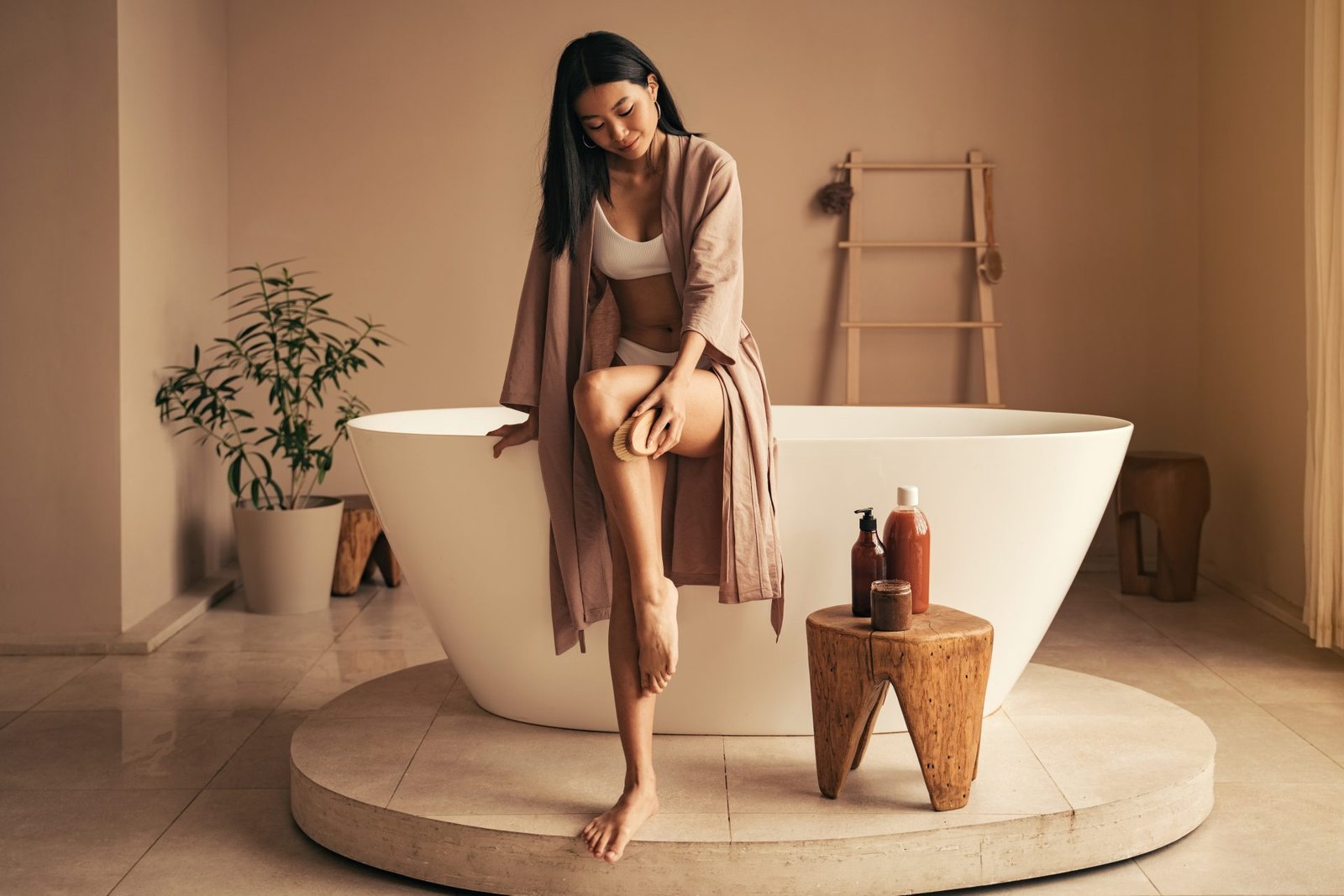 Giovane donna asiatica che mostra spazzolatutra a secco come si fa e massaggia le gambe con una spazzola di legno a secco in un lussuoso bagno accanto a una vasca da bagno in ceramica. Trattamento anticellulite.