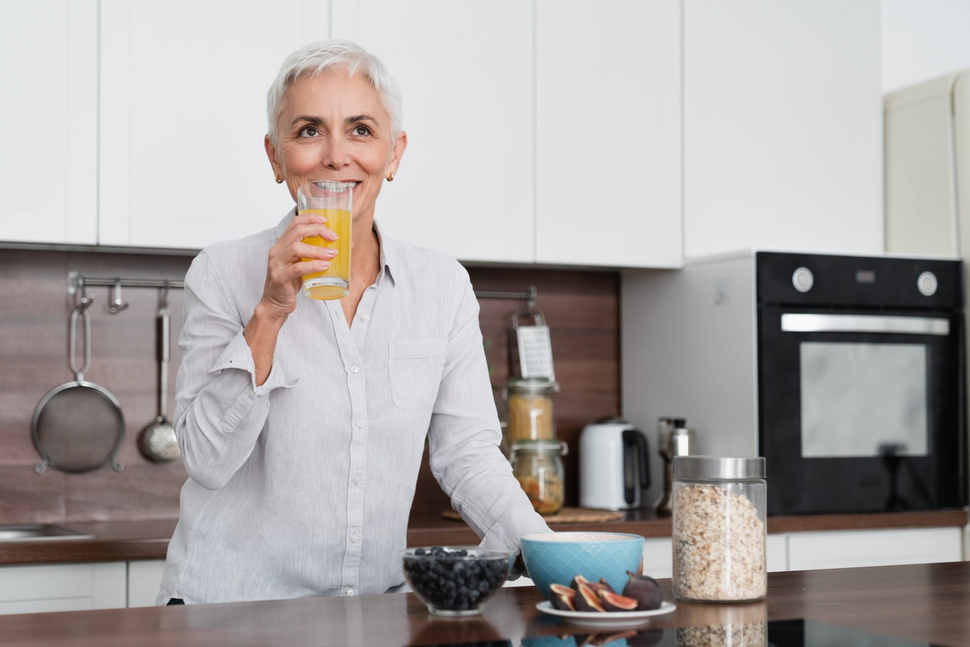 Donna matura in forma che beve succo d'arancia con vitamine mentre fa colazione nella cucina di casa e mangia granola sana con frutta e fiocchi d'avena. Colazione ideale in menopausa 