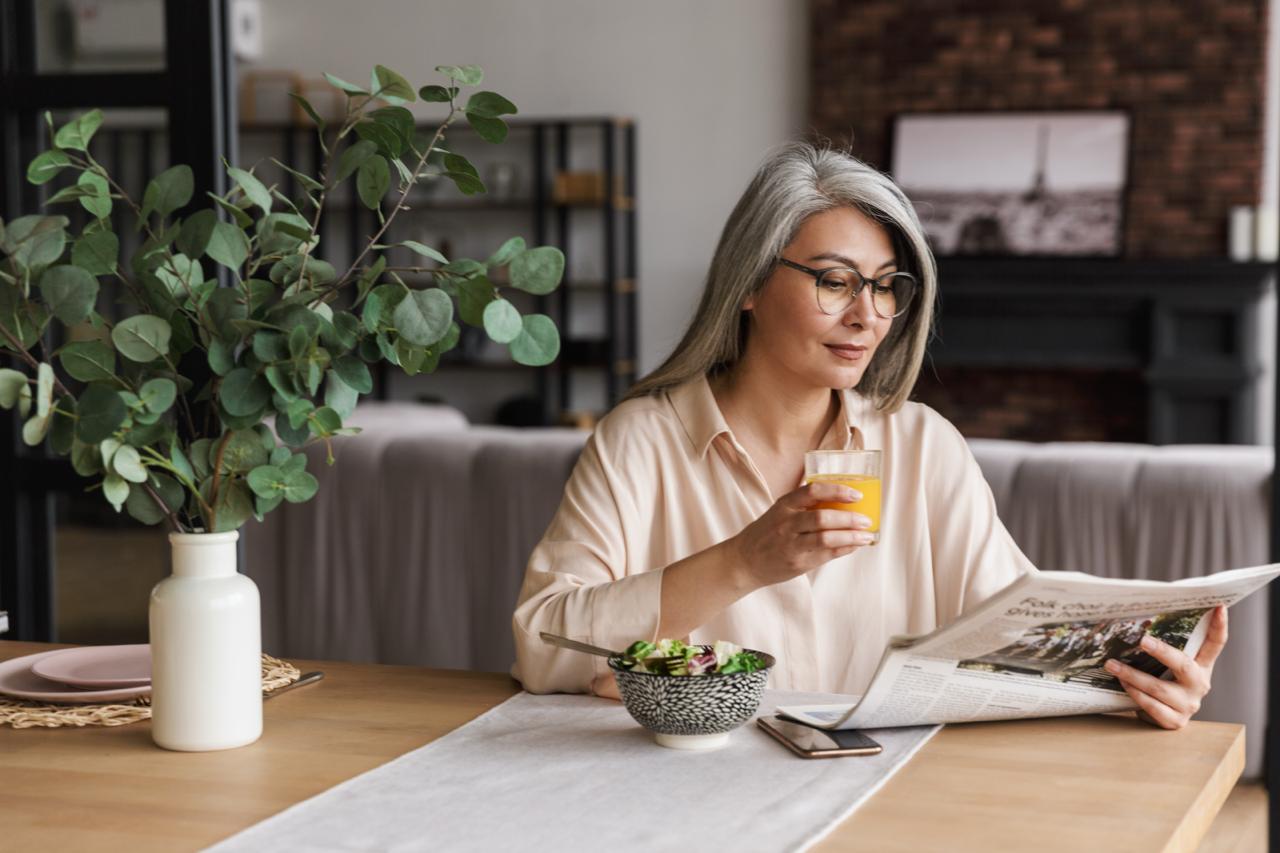 COLAZIONE IDEALE IN MENOPAUSA Donna concentrata che legge il giornale mentre beve succo DI FRUTTA CON CIOTOLA DELLA COLAZIONE DAVANTI