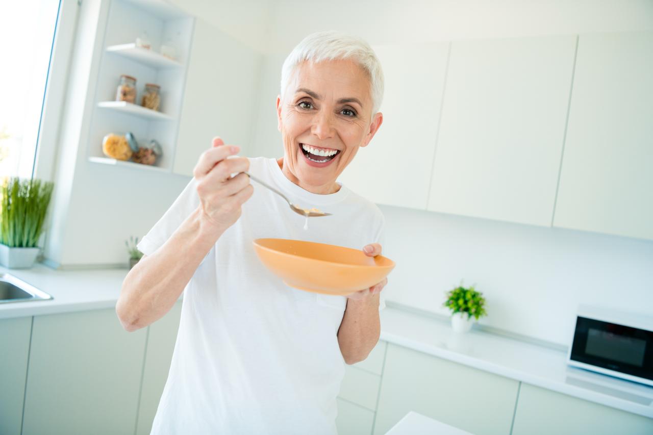 Una felice donna di mezza età con i capelli cortisorride in una cucina luminosa, tenendo in mano una ciotola e un cucchiaio- colazione ideale in menopausa