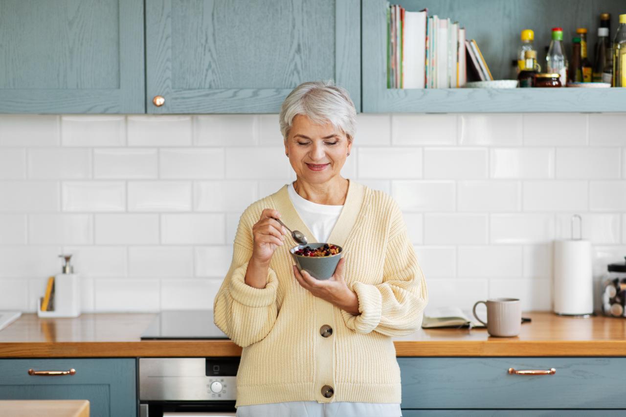 donna sorridente e felice che mangia cereali con un cucchiaio in cucina a casa CONCETTO DI COLAZIONE IDEALE IN MENOPAUSA