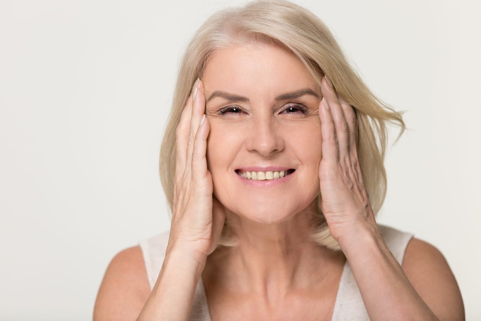 Donna anziana matura felice che tocca il viso con la pelle sana, morbida e idratata, isolata su sfondo grigio da studio, donna anziana di mezza età sorridente, concetto di trattamento di bellezza naturale anti-invecchiamento, ritratto