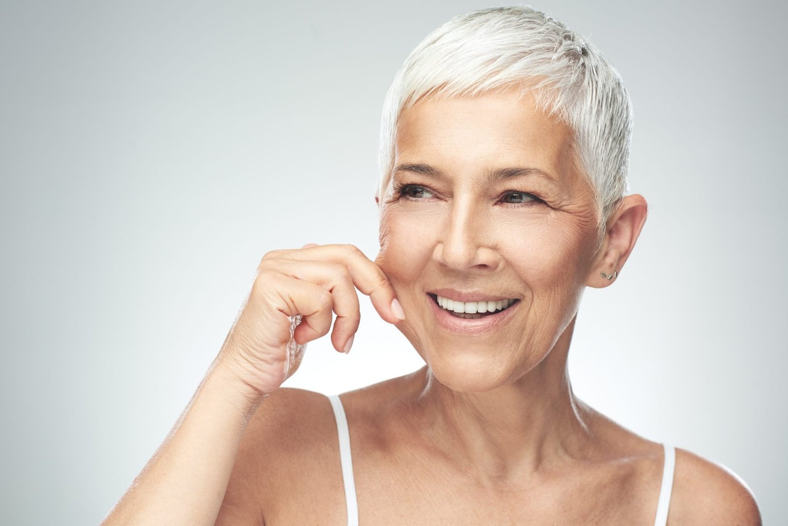 Splendida donna caucasica anziana e sorridente, con corti capelli grigi che si pizzicano la guancia. Fotografia di bellezza a tema skincare routine pelle matura