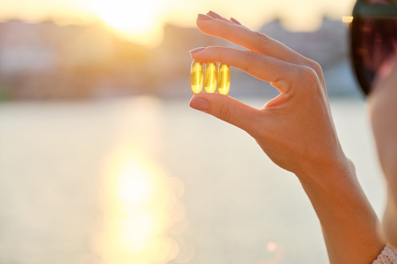 Capsula di gel giallo contenente vitamina D e omega-3 nella mano di una donna, sullo sfondo sole e mare.- CONCETTO DI OMEGA 3 QUANDO ASSUMERLI