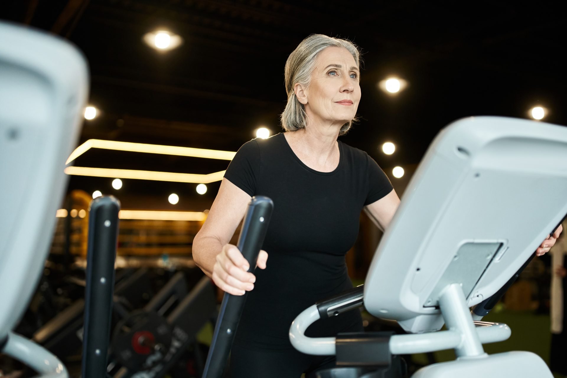 Una bella e allegra sportiva matura in maglietta nera si allena attivamente su un'ellittica. allenamento in menopausa 