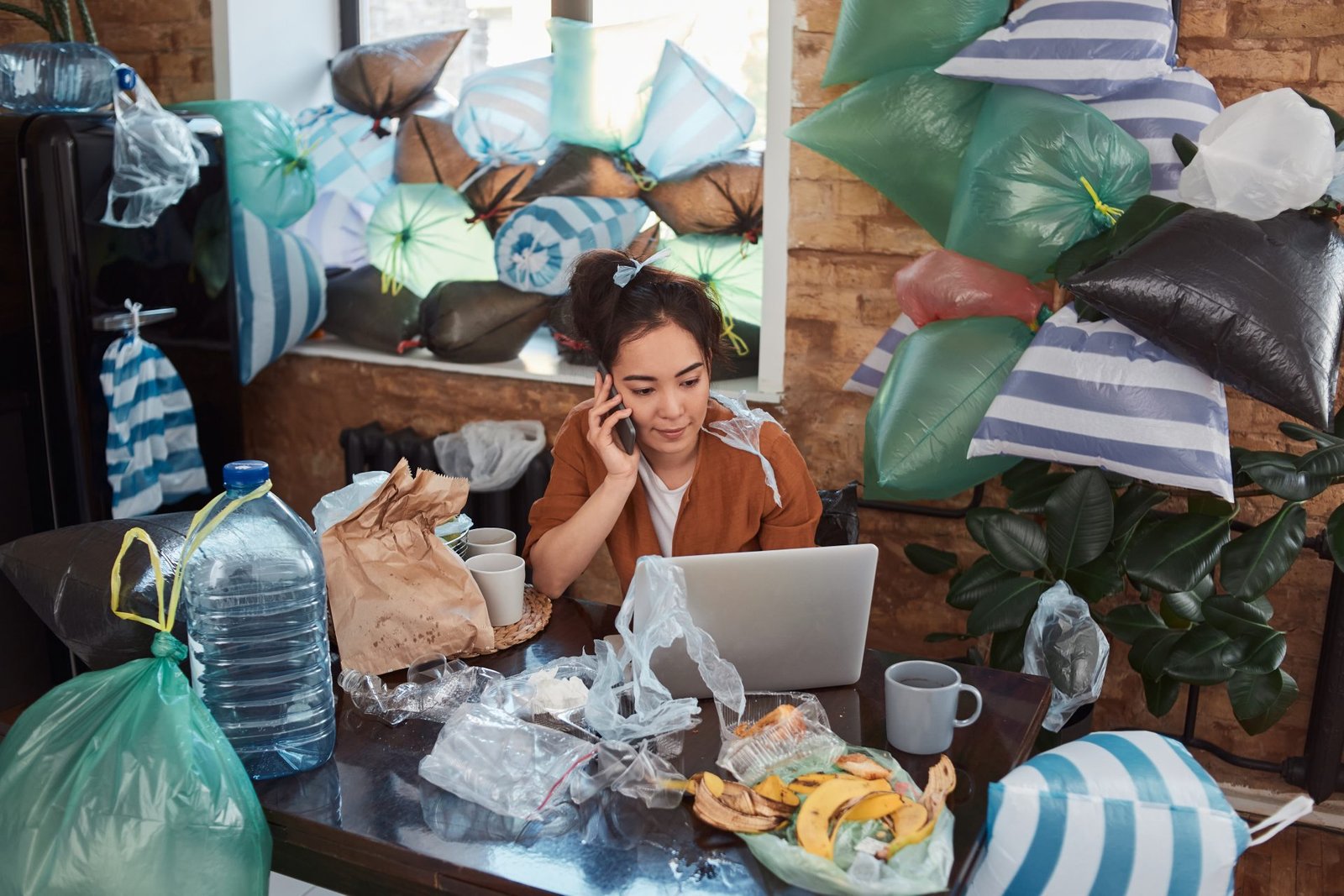 Una giovane donna comunica al telefono davanti a un computer portatile nel suo appartamento disordinato e disordinato, pieno di spazzatura ovunque - concetto di decluttering di primavera 